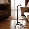 A black guitar stand is shown in a living room setting, with sofas and a rug visible. Náhľad obrázku produktu