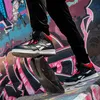 A person wearing black pants and Reebok high-top sneakers is standing on a piece of wood with graffiti in the background. Náhľad obrázku produktu