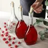 Three glass bottles with cork stoppers, two filled with red liquid and one empty, are arranged on a wooden surface with raspberries scattered around. A hand is shown inserting a cork into one of the bottles. Voir les images du produit