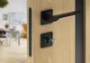 A close-up of a modern black door handle and lock mechanism on a light wood door, with a blurred interior room visible through the open doorway. Produktbild-Vorschau