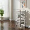 A multi-tiered rolling cart with wire baskets and a wooden top sits in a room with a plant and a painting on the wall. Voir les images du produit