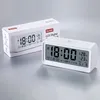 A white digital alarm clock is displayed next to its packaging on a light gray surface. Produktbild-Vorschau
