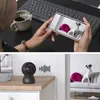 A split image showing a person using a smartphone to view a security camera feed, and a separate shot of the security camera and the room it's monitoring. Produktbild-Vorschau