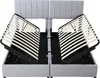 A grey upholstered bed frame with the slatted base lifted on both sides, revealing storage space underneath. Produktbild-Vorschau