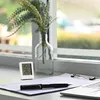 A digital thermometer and hygrometer sits on a desk next to a document, pen, and glasses, with a plant in a glass vase in the background, near a window. Anteprima dell’immagine prodotto