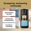 A close-up shot of a black Tchibo Qbo coffee machine with a blue accent, brewing espresso into a glass, with text overlays describing its features. The background is blurred with hints of pink and blue. Produktbild-Vorschau