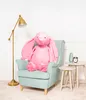 A large pink plush bunny sits in a light blue armchair next to two small side tables. A floor lamp is to the left of the armchair. The background is a white wall with decorative molding. Produktbild-Vorschau