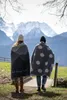 Two people wrapped in blankets stand on a path with mountains in the background. Produktbild-Vorschau