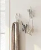 Three wall hooks shaped like climbing figures are mounted on a white wall. One hook is holding a folded plaid scarf. Another hook has a necklace hanging from it. The third hook is empty. Náhled obrázku produktu