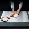 A person's hands are kneading dough on a stainless steel surface, with eggs and flour nearby. Produktbild-Vorschau