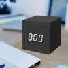 A black digital alarm clock with white LED display showing 8:00 is placed on a wooden desk next to a white object, possibly a power bank or book. A laptop is visible in the background. Produktbild-Vorschau