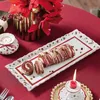 A festive table setting with a Christmas-themed cake plate, a roulade cake, flowers, and candles. Náhľad obrázku produktu
