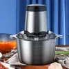A stainless steel electric meat grinder with a clear lid sits on a wooden table, with a blue curtain in the background. Ingredients like raw meat, garlic, and chopped vegetables are visible. Produktbild-Vorschau