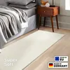 A cream-colored shaggy rug is placed on a wooden floor next to a bed and a nightstand. Náhled obrázku produktu