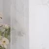 A close-up view of a sheer curtain with a gold lotus pattern, with a vase of white flowers in the foreground. Produktbild-Vorschau