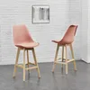 Two pink bar stools with wooden legs are shown in front of a white brick wall with a painting. Náhled obrázku produktu