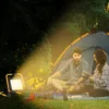 A couple is camping next to a tent, illuminated by a bright work light. Produktbild-Vorschau