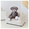 A large gray teddy bear is sitting on a cream-colored armchair in a bright room. Podgląd zdjęcia produktu