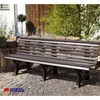 A brown garden bench sits on a cobblestone patio in front of a stone wall and greenery. Produktbild-Vorschau