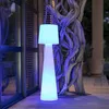 A tall, cone-shaped floor lamp with a pleated shade is illuminated with blue and white light, standing on a wooden deck next to a large, gnarled tree trunk. Produktbild-Vorschau