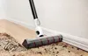 A cordless vacuum cleaner is shown cleaning a wooden floor near a white wall and baseboard. Dirt and debris are visible on the floor. Produktbild-Vorschau