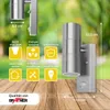 A stainless steel wall lamp with informational icons and dimensions displayed against a blurred green and wooden background. Produktbild-Vorschau
