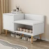 A white shoe cabinet with a cushioned bench and storage shelves, featuring gold metal legs, placed against a wall with a curtain. Produktbild-Vorschau