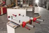 A workshop setting with a person working in the background and a workbench with clamps holding a piece of material in the foreground. Produktbild-Vorschau