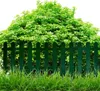 A dark green picket fence in front of a lush green bush, with grass in the foreground. Produktbild-Vorschau