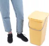 A person's legs and feet are shown standing next to a yellow trash can with a lid and handle. Podgląd zdjęcia produktu