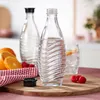 Two glass bottles of sparkling water, one with a black cap, are displayed on a wooden board with oranges and raspberries. A glass of iced drink is in the background. Produktbild-Vorschau