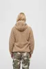 A woman is shown from behind wearing a tan teddy fleece jacket and camouflage pants. Produktbild-Vorschau