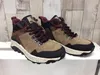 A pair of beige and brown hiking boots are displayed on a white surface against a wood-paneled background. Produktbild-Vorschau