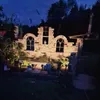 A nighttime garden scene featuring a decorative brick structure resembling ancient ruins with arched windows, illuminated by solar lights. Produktbild-Vorschau