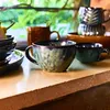 Several ceramic mugs and bowls are arranged on a textured surface, with a blurred background of greenery visible through a window. Podgląd zdjęcia produktu