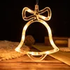 A close-up of a clear bell-shaped LED light with a bow on top, illuminated with warm yellow lights, hanging against a dark background. The bell is made of clear plastic and contains small LED lights. In the background, there are some decorative items like  Produktbild-Vorschau