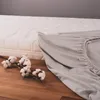 A light gray fitted sheet is partially laid out next to a mattress, with cotton sprigs on a wooden surface in the foreground. Náhled obrázku produktu