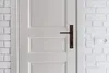 A close-up view of a white door with a bronze door handle and a white brick wall with an electrical outlet to the right. Náhled obrázku produktu