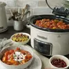 A Crockpot slow cooker is shown on a kitchen counter, surrounded by bowls of food and kitchen utensils. Produktbild-Vorschau