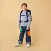 A young girl wearing a backpack and colorful jeans stands against a plain background. Anteprima dell’immagine prodotto