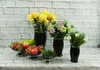 A still life arrangement of flowers and fruit in black vases and bowls, set against a white brick wall. Produktbild-Vorschau