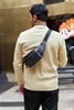 A man wearing a beige sweater and black pants is seen from behind, with a black fanny pack worn across his chest. Voir les images du produit