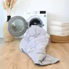 A gray comforter is being pulled out of a washing machine, with a decorative vase and a laundry basket in the background. Produktbild-Vorschau