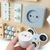 A hand is shown holding a wooden plug with two prongs, about to insert it into a socket on a wooden Montessori busy board. Other features of the busy board, such as beads and elastic strings, are visible. Produktbild-Vorschau
