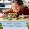 A young girl is playing with a LEGO Minecraft set on a wooden table. The set includes a stable structure, various Minecraft characters, and animals. The background shows a blurred indoor environment. Podgląd zdjęcia produktu
