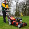 A man is pushing a red and black lawnmower on a grassy lawn with trees in the background. Produktbild-Vorschau