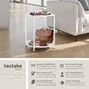 A white round side table with two shelves, placed in a modern living room setting next to a sofa. The table has books, a briefcase, and a cup on it. The image also includes marketing text and icons describing the product's features. Náhled obrázku produktu