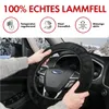A person's hands are holding a black sheepskin steering wheel cover inside a car, with a snowy road visible through the windshield. Produktbild-Vorschau