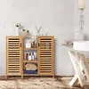 A bamboo bathroom cabinet with slatted doors and open shelves, displaying various bathroom items and folded towels, against a tiled wall and wooden floor. Produktbild-Vorschau