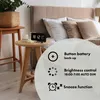 A digital alarm clock is shown on a bedside table next to a bed. The image includes text overlays highlighting product features. Produktbild-Vorschau